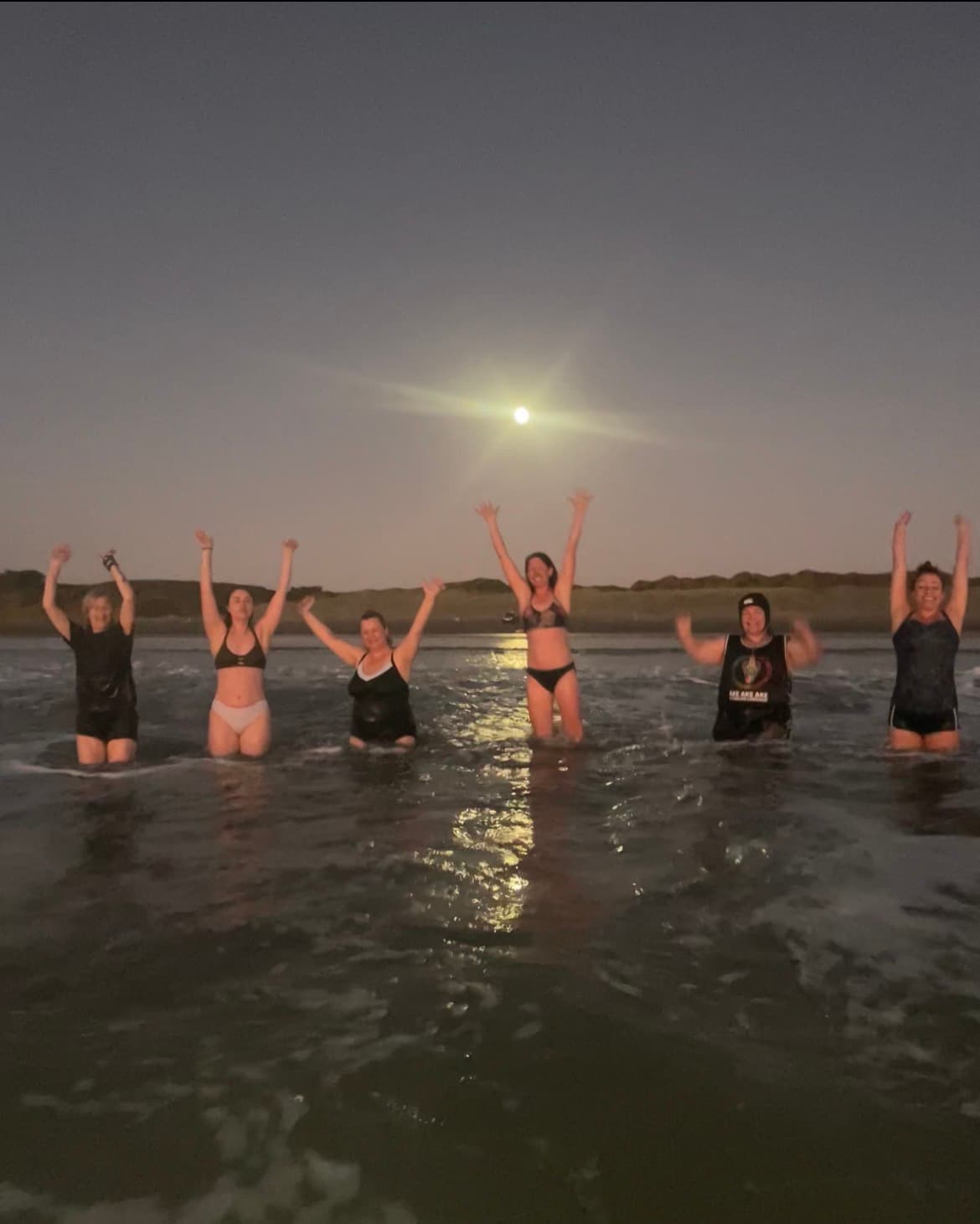 Six women with arms raised stand in the ocean under a bright full moon.